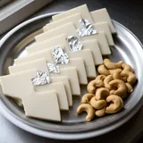 Diamond-shaped kaju katli pieces, some with silver leaf, beside raw cashews