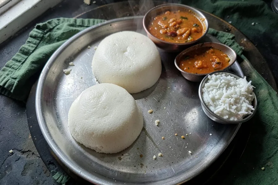 Two steamed idli on a steel plate with sambar and coconut chutney