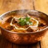 Rich mutton curry in a copper pot with cilantro garnish.
